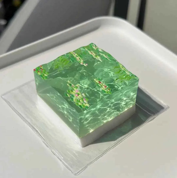Green translucent cube squishy with colorful sprinkles, resting on white tray, soft and slow-rising.