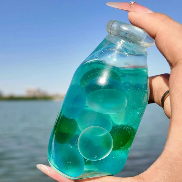 Hand holding a transparent bottle-shaped squishy filled with blue-green liquid and round beads, part of Lovesquishy's Crystal Bottle Series.